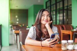 réfléchi Jeune asiatique femme portant robe séance à une restaurant pour une petit-déjeuner, en portant une téléphone intelligent tandis que étayer sa menton photo