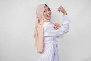 excité asiatique musulman femme portant blanc robe et hijab souriant en toute confiance tandis que posant fort geste par levage sa bras et muscles. Ramadan et eid mubarak concept photo