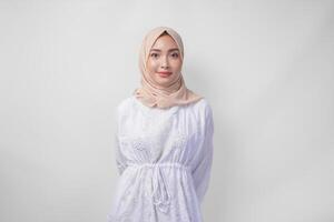 attrayant asiatique musulman femme dans blanc robe et hijab souriant à le caméra tandis que permanent officiellement plus de isolé blanc Contexte photo