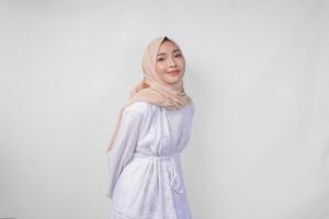 attrayant asiatique musulman femme dans blanc robe et hijab souriant à le caméra tandis que permanent officiellement plus de isolé blanc Contexte photo