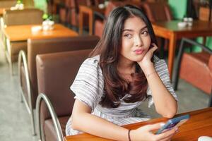 réfléchi Jeune asiatique femme portant robe séance à une restaurant pour une petit-déjeuner, en portant une téléphone intelligent tandis que étayer sa menton photo