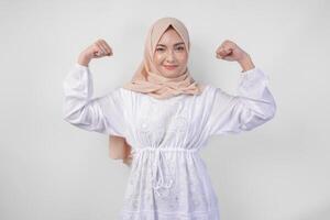 excité asiatique musulman femme portant blanc robe et hijab souriant en toute confiance tandis que posant fort geste par levage sa bras et muscles. Ramadan et eid mubarak concept photo