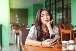 réfléchi Jeune asiatique femme portant robe séance à une restaurant pour une petit-déjeuner, en portant une téléphone intelligent tandis que étayer sa menton photo