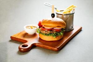 classique cajun du boeuf Burger comprendre fromage tranche, tomate, oignon et salade feuille servi sur en bois planche avec tremper et français frites seau isolé sur gris Contexte côté vue de apéritif vite nourriture photo