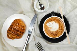 chaud macchiato café latté art servi dans tasse avec croissant, bouffée Pâtisserie, pain et couteau isolé sur serviette de table Haut vue café petit déjeuner photo