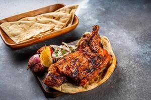 grillé poulet moitié un barbecue gril avec Pita pain, Nan, tandoori rôti, raita, tremper et salade, tomate oignon et citron servi dans en bois planche Haut vue sur Contexte épicé barbecue photo