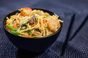 mixte frit nouilles avec avec poulet, carotte, printemps oignon, cloche poivre, Oeuf et baguettes servi dans bol côté vue de chinois nourriture photo