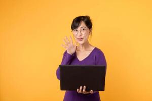 une portrait de une Jeune et sur de soi femme d'affaires portant une violet chemise et lunettes, en portant une ordinateur portable, signifiant sa compétence et connaissance dans le numérique monde contre une Jaune Contexte. photo