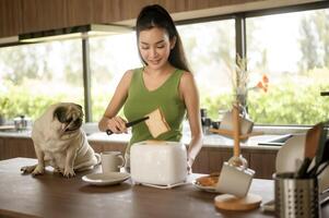 asiatique femme en train de préparer café et pain grillé pain pour petit déjeuner prendre plaisir avec chien à le cuisine table dans le Matin photo