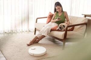 Jeune asiatique femme relaxant avec carlin chien dans vivant pièce tandis que robotique vide nettoyeur travail photo