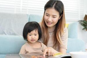 heureuse mère asiatique se détendre et lire un livre avec bébé temps ensemble à la maison. un parent s'assoit sur un canapé avec sa fille et lit une histoire. apprendre le développement, la garde d'enfants, le rire, l'éducation, la narration, la pratique. photo