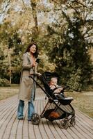 Jeune femme avec mignonne bébé fille dans bébé poussette en utilisant mobile téléphone à le l'automne parc photo