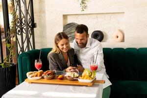 Jeune couple utilisant mobile téléphone tandis que ayant le déjeuner et en buvant Frais pressé jus dans le restaurant photo