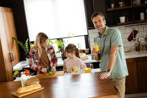 joyeux homme avec une verre de Orange jus tandis que une femme et enfant préparer nourriture dans une confortable cuisine photo