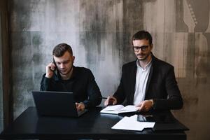 deux les hommes d'affaires dans un Bureau souriant à le caméra tandis que travail ensemble derrière une portable ordinateur. photo