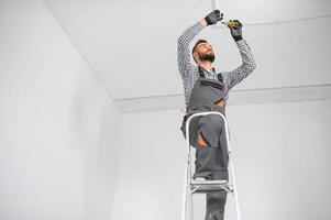 dépanneur ou professionnel électricien dans vêtements de travail installation lumière taches, permanent sur le échelle dans le blanc vivant pièce photo