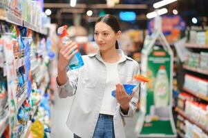 intéressé Jeune femme fabrication achats dans Ménage produits chimiques magasin, en train de lire Étiquettes sur bouteilles photo