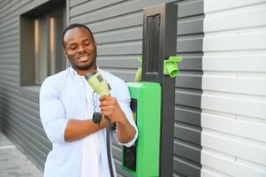 portrait de une Jeune homme permanent avec mise en charge câble près le mise en charge gare. concept de vite Accueil voiture chargeurs. photo