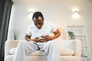 africain américain gars ayant estomac mal après en mangeant émouvant douloureux estomac Souffrance de douleur séance sur canapé à maison. photo