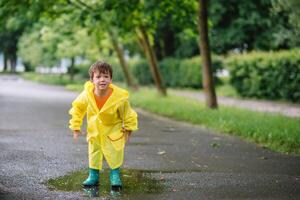 peu garçon en jouant dans pluvieux été parc. enfant avec parapluie, imperméable manteau et bottes sauter dans flaque et boue dans le pluie. enfant en marchant dans été pluie Extérieur amusement par tout temps. content enfance photo