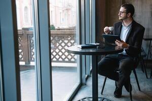 Beau homme d'affaire en utilisant une numérique tablette et en buvant café tandis que séance dans café dans le ville centre. photo