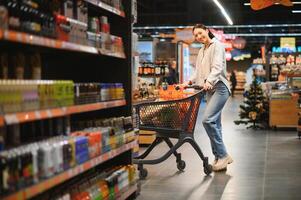 souriant content femme profiter achats à le supermarché, elle est penché sur une plein Chariot photo