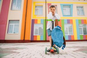 adolescent garçon avec donner un coup scooter près moderne école. enfant avec sac à dos et livre en plein air. début de cours. premier journée de automne. retour à école. photo