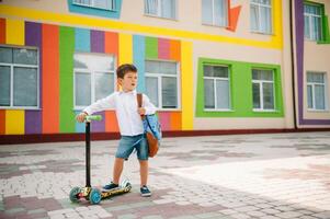 adolescent garçon avec donner un coup scooter près moderne école. enfant avec sac à dos et livre en plein air. début de cours. premier journée de automne. retour à école. photo