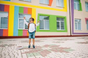 retour à école. content souriant garçon dans des lunettes est Aller à école pour le premier temps. enfant avec sac à dos et livre en plein air. début de cours. premier journée de tomber photo