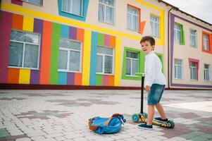 adolescent garçon avec donner un coup scooter près moderne école. enfant avec sac à dos et livre en plein air. début de cours. premier journée de automne. retour à école. photo