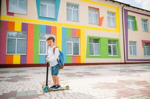adolescent garçon avec donner un coup scooter près moderne école. enfant avec sac à dos et livre en plein air. début de cours. premier journée de automne. retour à école. photo