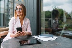 portrait de une charmant affaires femme bavardage sur sa intelligent téléphone tandis que attendre Quelqu'un dans café en plein air, magnifique femelle en utilisant mobile téléphone tandis que séance dans confortable restaurant dans le Frais air pendant photo