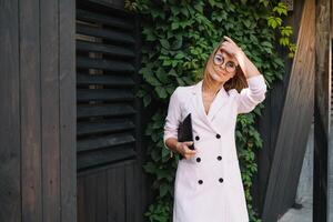 portrait de une réussi femme d'affaires en portant une numérique tablette tampon et une dossier tandis que fièrement permanent près bureau. photo