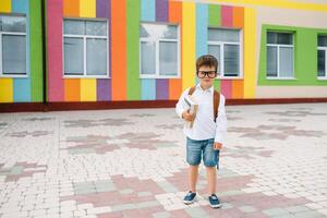 retour à école. content souriant garçon dans des lunettes est Aller à école pour le premier temps. enfant avec sac à dos et livre en plein air. début de cours. premier journée de tomber photo