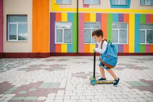 adolescent garçon avec donner un coup scooter près moderne école. enfant avec sac à dos et livre en plein air. début de cours. premier journée de automne. retour à école. photo