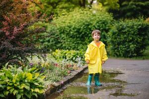 peu garçon en jouant dans pluvieux été parc. enfant avec parapluie, imperméable manteau et bottes sauter dans flaque et boue dans le pluie. enfant en marchant dans été pluie Extérieur amusement par tout temps. content enfance. photo