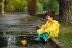 enfant en jouant avec jouet bateau dans flaque. enfant jouer Extérieur par pluie. tomber pluvieux temps en plein air activité pour Jeune les enfants. enfant sauter dans boueux flaques d'eau. imperméable veste et bottes pour bébé. enfance photo