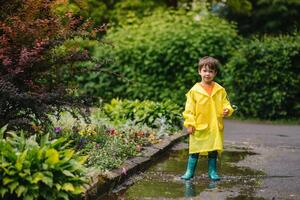 peu garçon en jouant dans pluvieux été parc. enfant avec parapluie, imperméable manteau et bottes sauter dans flaque et boue dans le pluie. enfant en marchant dans été pluie Extérieur amusement par tout temps. content enfance. photo