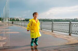 peu garçon en jouant dans pluvieux été parc. enfant avec parapluie, imperméable manteau et bottes sauter dans flaque et boue dans le pluie. enfant en marchant dans été pluie Extérieur amusement par tout temps. content enfance. photo