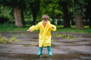 peu garçon en jouant dans pluvieux été parc. enfant avec parapluie, imperméable manteau et bottes sauter dans flaque et boue dans le pluie. enfant en marchant dans été pluie Extérieur amusement par tout temps. content enfance photo