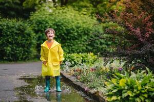 peu garçon en jouant dans pluvieux été parc. enfant avec parapluie, imperméable manteau et bottes sauter dans flaque et boue dans le pluie. enfant en marchant dans été pluie Extérieur amusement par tout temps. content enfance. photo