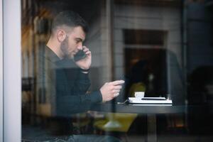 Jeune homme d'affaire parlant sur mobile téléphone tandis que travail sur portable dans café. photo