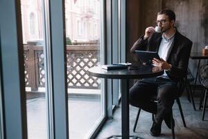 Beau homme d'affaire en utilisant une numérique tablette et en buvant café tandis que séance dans café dans le ville centre. photo