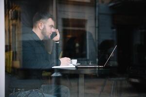 Jeune homme d'affaire parlant sur mobile téléphone tandis que travail sur portable dans café. photo