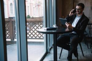 Jeune homme d'affaire parlant sur mobile téléphone tandis que travail sur tablette dans café. photo