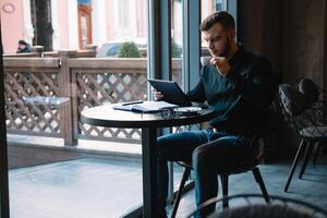 Beau homme d'affaire en utilisant une numérique tablette et en buvant café tandis que séance dans café dans le ville centre. photo