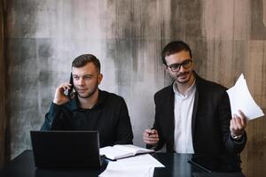 deux les hommes d'affaires dans un Bureau souriant à le caméra tandis que travail ensemble derrière une portable ordinateur. photo