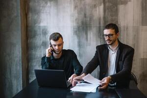 deux les hommes d'affaires dans un Bureau souriant à le caméra tandis que travail ensemble derrière une portable ordinateur photo