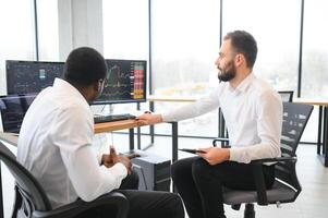 deux diverse collègues les commerçants parlant à chaque autre, séance dans le Bureau dans de face de plusieurs ordinateur écrans. Stock commerce, personnes, affaires concept photo