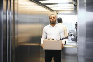 une Jeune africain homme mis à la porte de travail est permanent dans le ascenseur avec une boîte de des choses photo